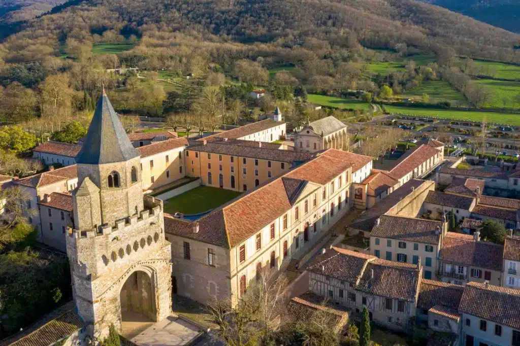 Abbaye historique : Tour fortifiée et bâtiments ocre. Vue aérienne d'une abbaye historique. Tour d'entrée médiévale, clocher pointu, longs bâtiments ocre, toits en tuiles rouges. Vallée et collines boisées.