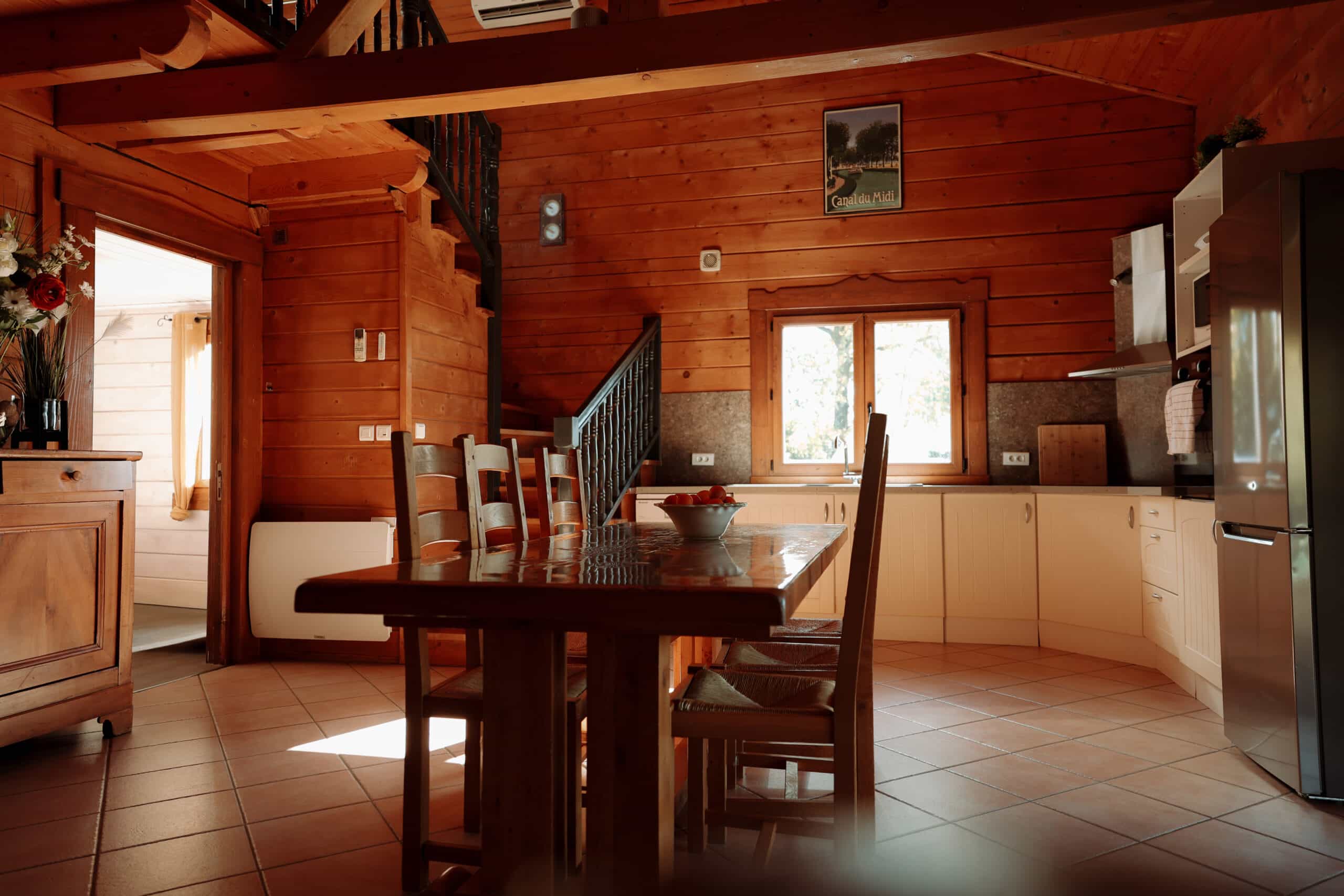 Intérieur chalet bois, salle à manger et cuisine. Table lustrée, chaises rustiques, escalier sombre. Sol terracotta. Lumière du jour. Affiche "Canal du Midi".