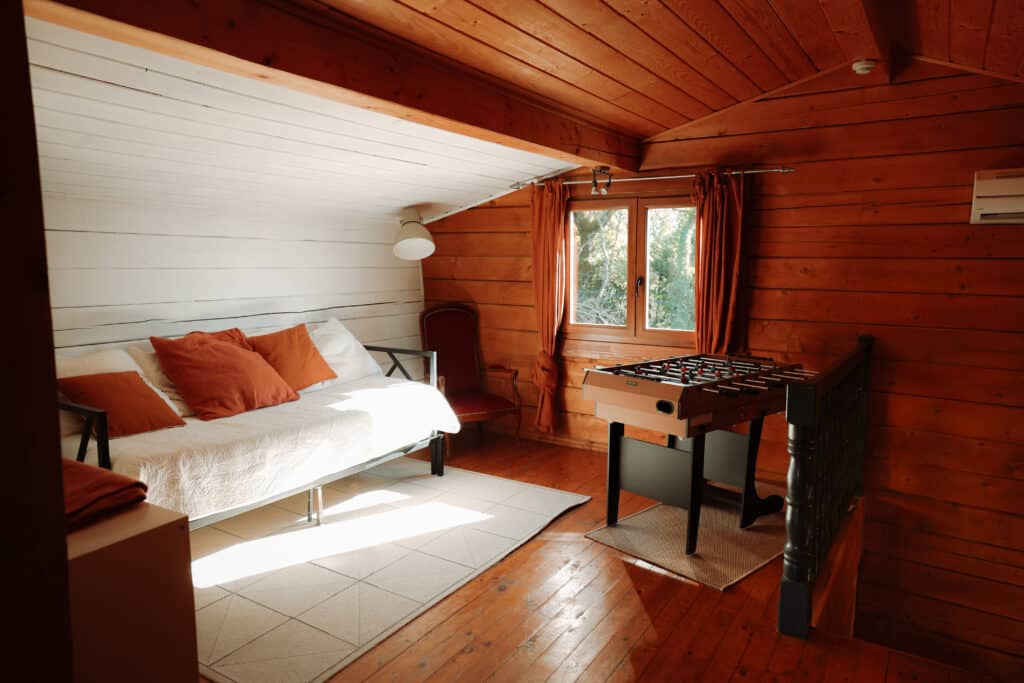 Loft en chalet bois : coin détente lumineux avec foosball. Intérieur de chalet lumineux. Plafond et murs en bois brun. Canapé blanc, oreillers orange, et table de foosball près de la fenêtre.