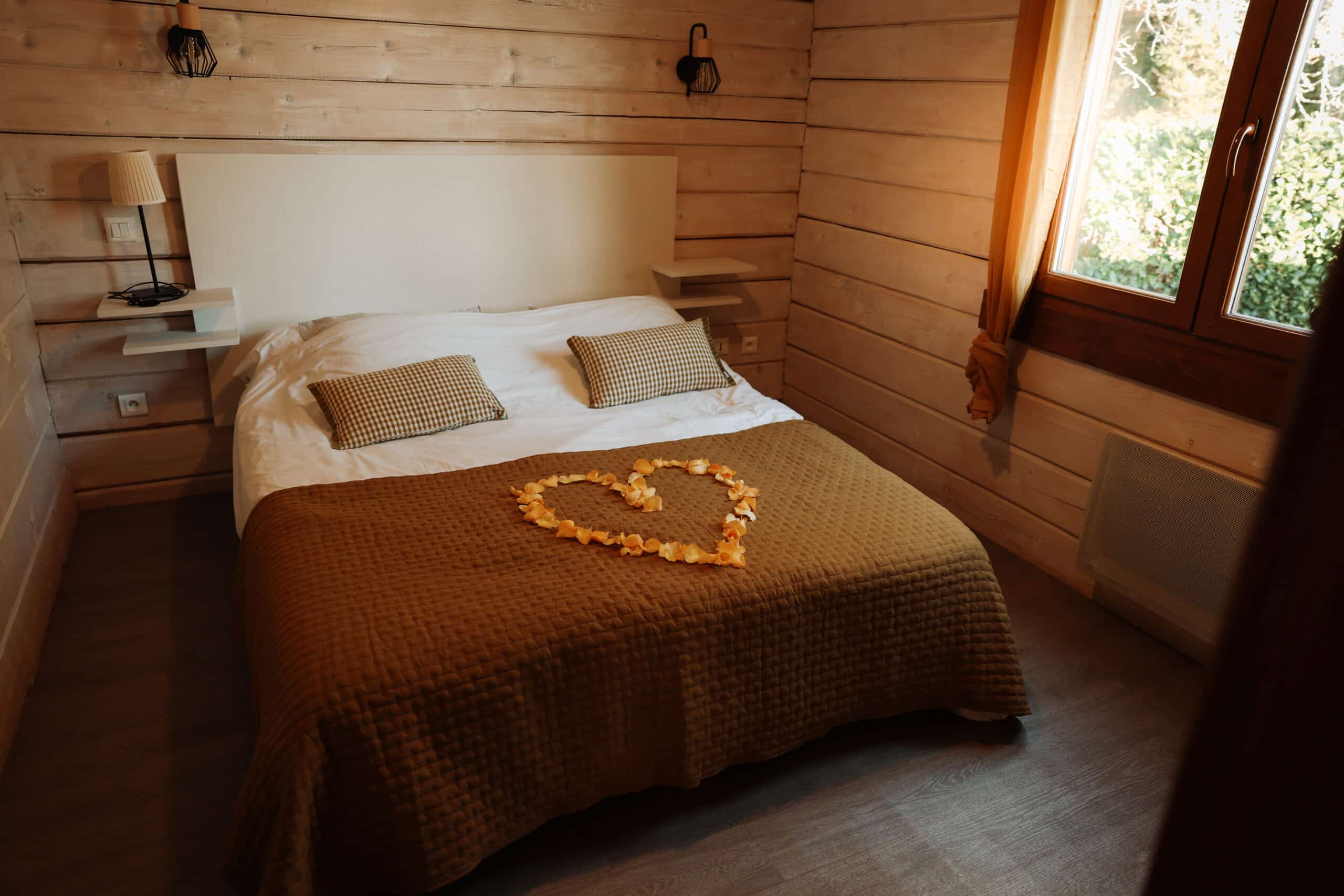 Lit double décoré d'un cœur de pétales de rose dans une chambre romantique en bois. Murs de cabane et grande fenêtre lumineuse.