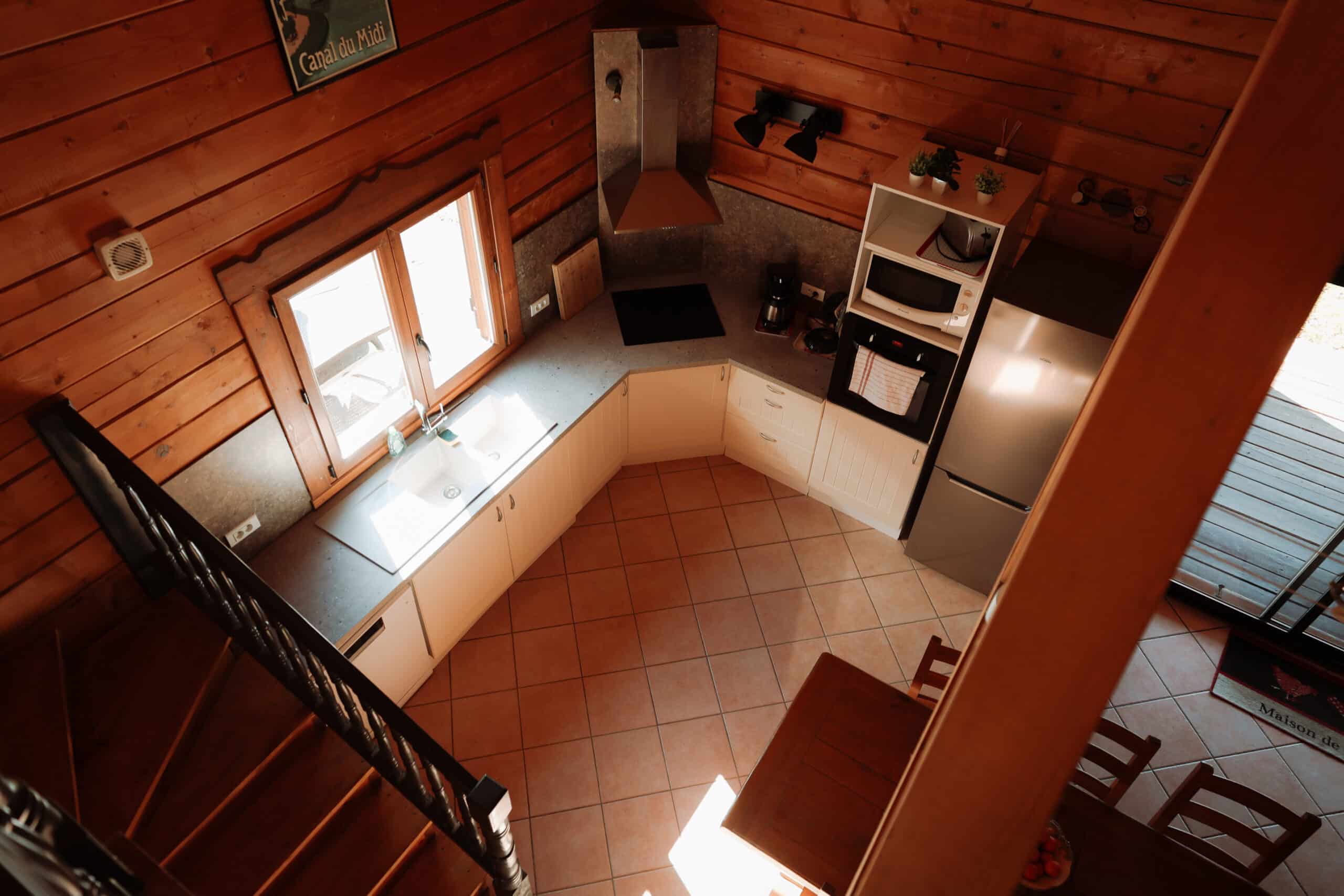 Cuisine de chalet rustique, vue plongeante. Murs en bois, carrelage terracotta. Évier sous fenêtre, électros inox. Affiche Canal du Midi.