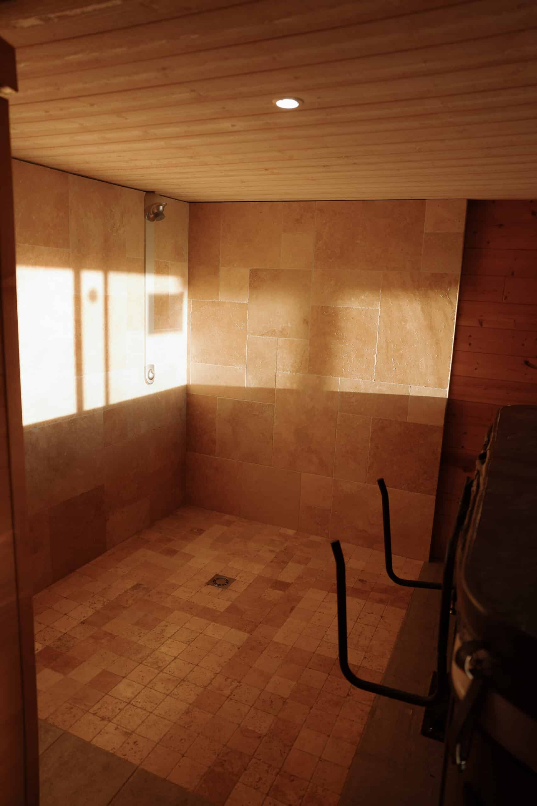 Grande douche italienne en travertin et bois avec lumière naturelle intense. Sol carrelé, plafonds lambrissés, barres de soutien noires.