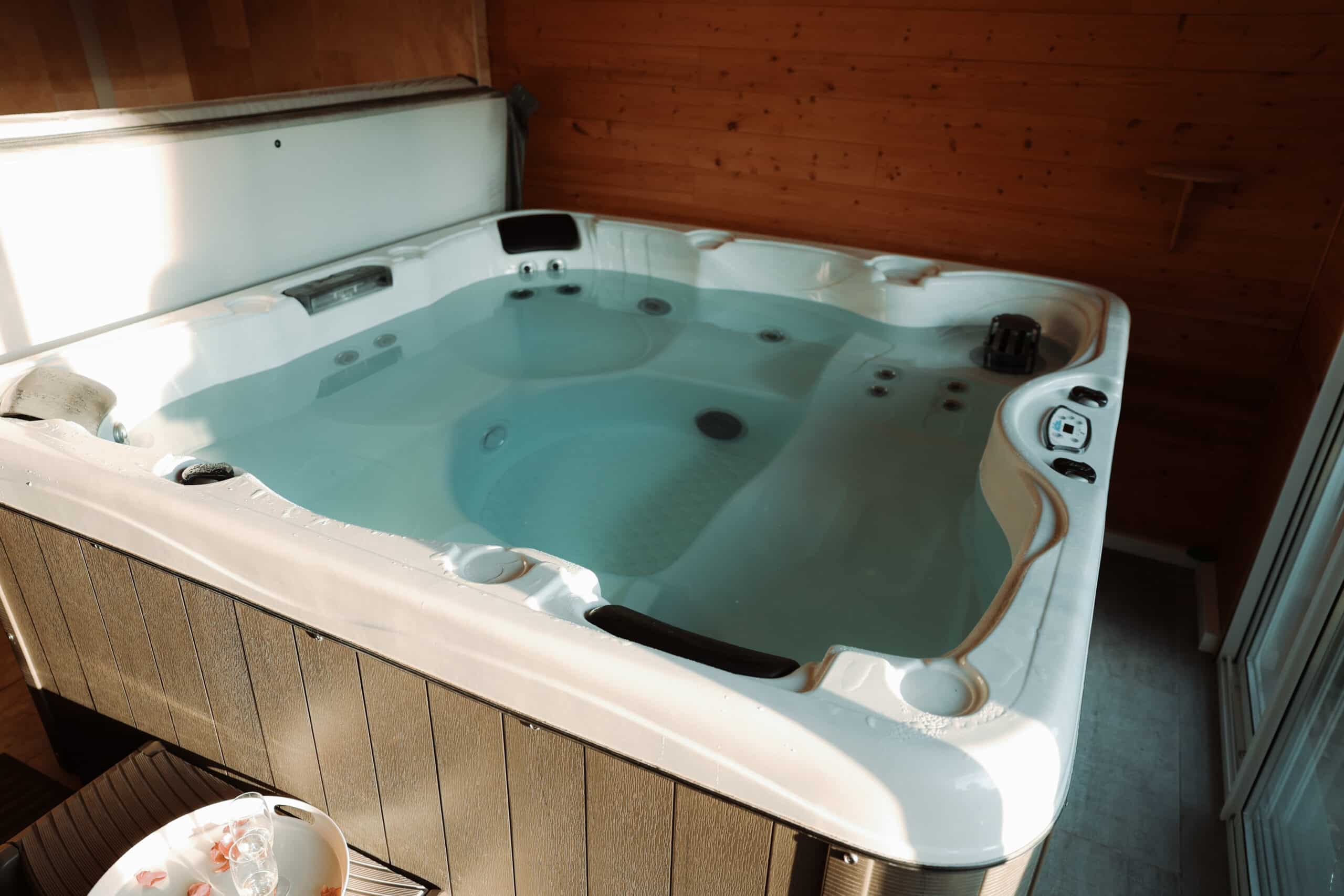 Grand spa intérieur en coque blanche et jupes en bois, rempli d'eau. La lumière du soleil éclaire la zone de détente et de luxe.