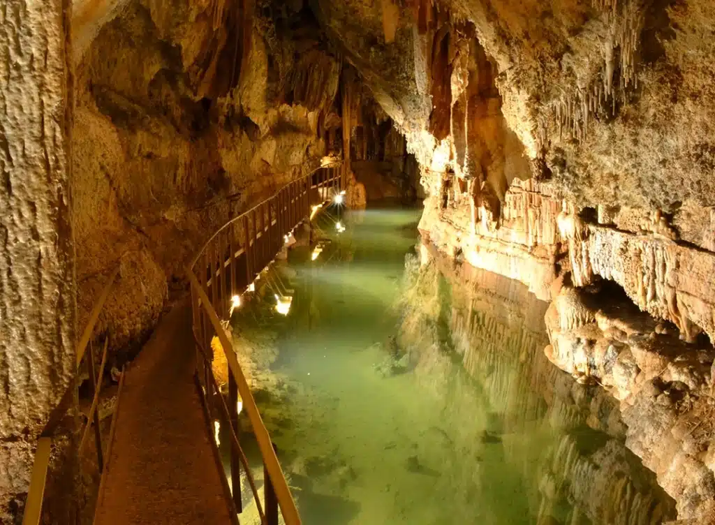 Grotte : Rivière souterraine et passerelle éclairée Passerelle métallique longeant une rivière souterraine aux eaux vertes dans une grotte éclairée. Stalactites et formations calcaires.