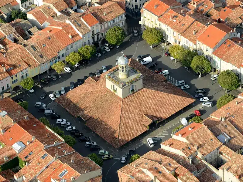 Place du village : halle et toits de tuiles, vue aérienne. Halle historique à coupole, entourée d'une place animée et de toits de tuiles rouges. Nombreux véhicules garés près des façades claires.
