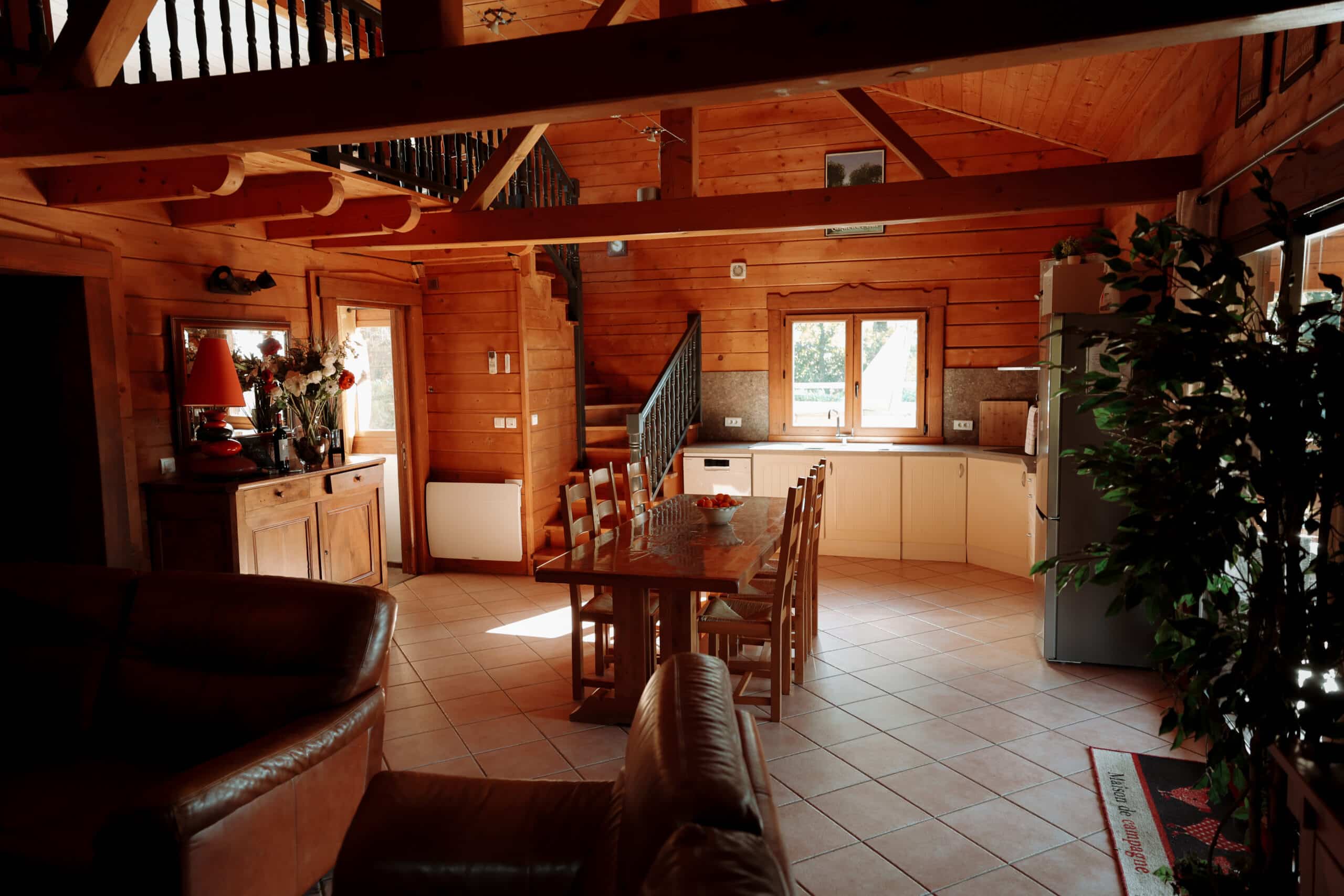 Intérieur chaleureux de chalet en bois rustique. Salon, salle à manger avec grande table, et cuisine claire. Escalier visible et poutres apparentes.