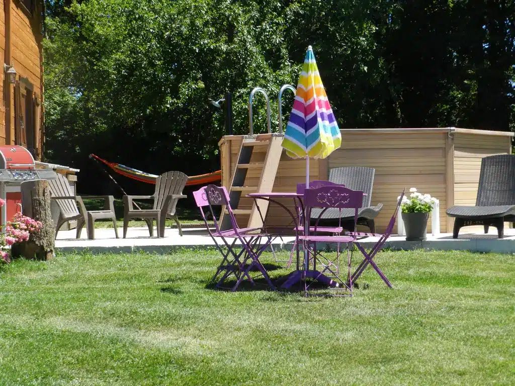 Jardin ensoleillé avec piscine hors-sol en bois, mobilier de jardin violet et parasol rayé. Barbecue et hamac pour la détente estivale.