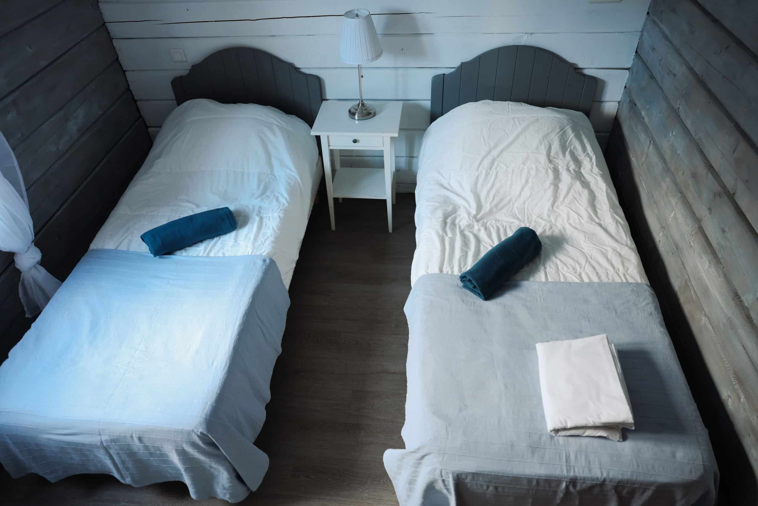 Chambre double rustique dans un chalet en bois. Deux lits jumeaux avec draps blancs, jetés bleus et serviettes roulées. Lampe sur table de nuit.