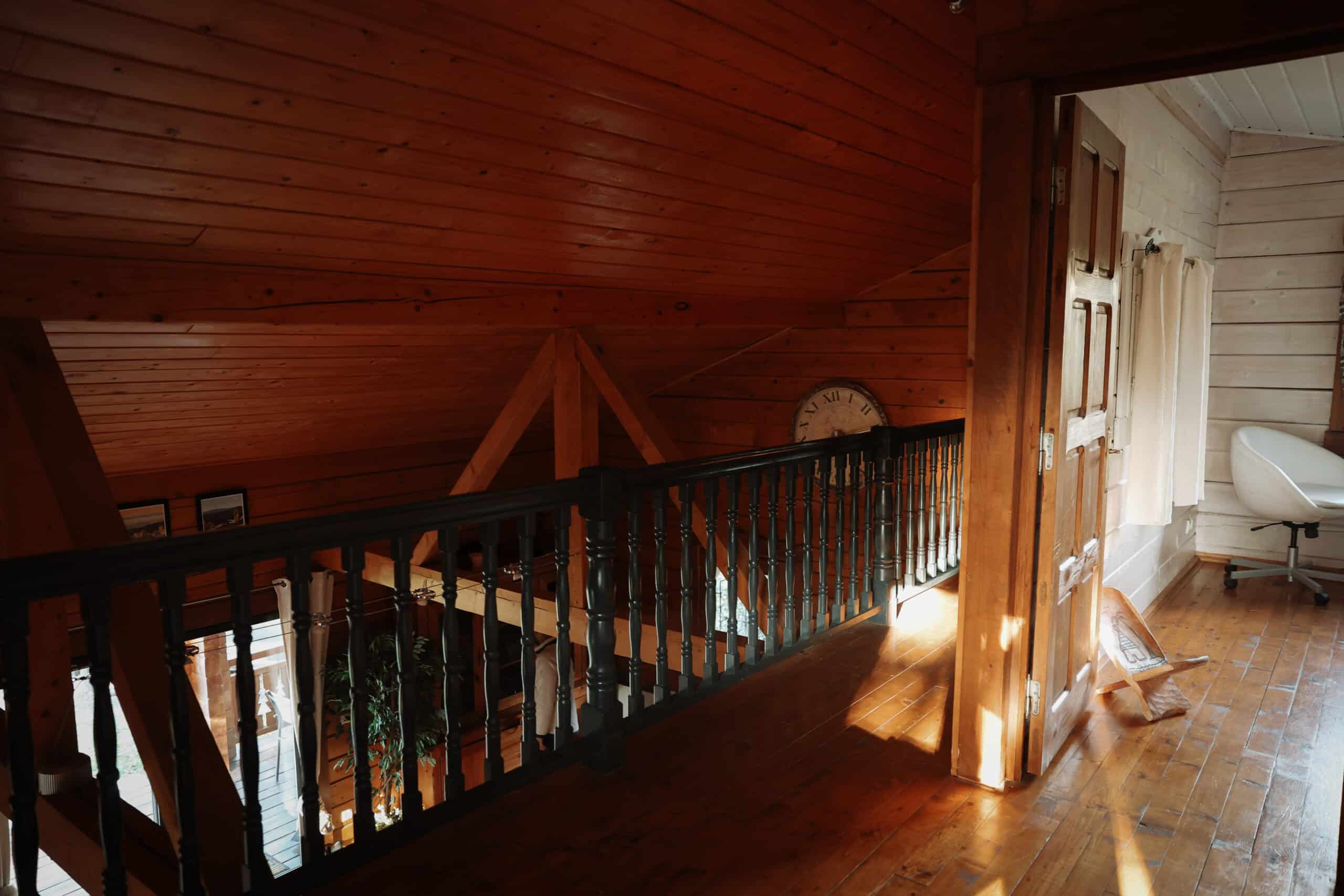 Intérieur de chalet en bois rouge. Mezzanine avec balustrade sculptée noire. Poutres apparentes, sol verni et grande horloge murale rustique.