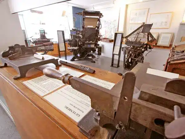 Musée de l'imprimerie : machines historiques et Gutenberg. Musée de l'imprimerie. Outils de composition en métal et textes "GUTENBERG" sur un comptoir. Deux grandes machines à imprimer historiques au fond.