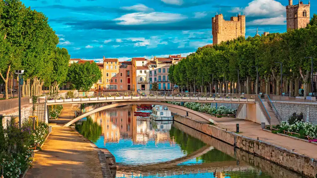 Perpignan : le canal de la Basse et ses reflets colorés. Perpignan : Canal de la Basse, reflet des maisons colorées et des platanes, pont et tours médiévales du Castillet sous ciel bleu.