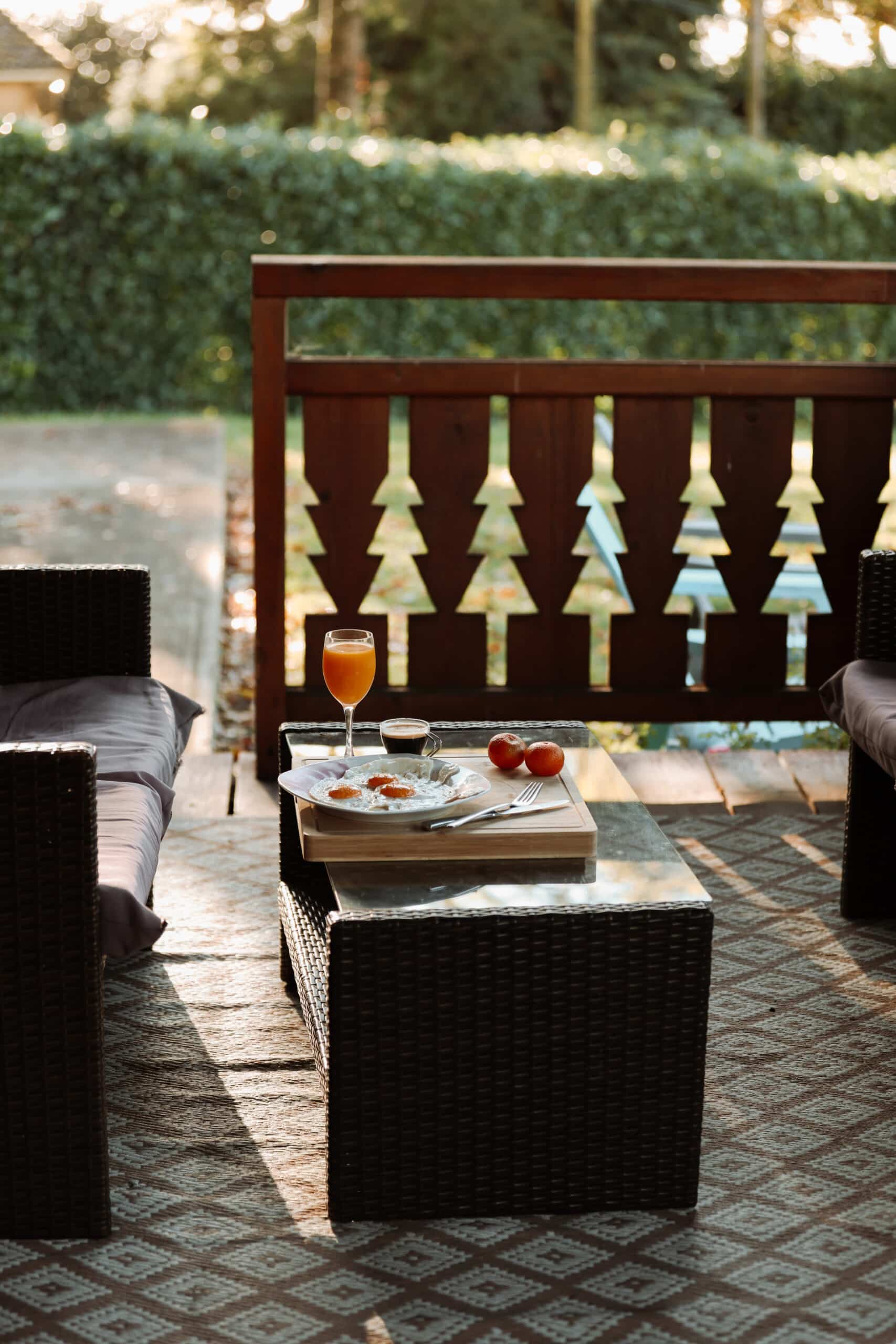 Petit déjeuner sur terrasse ensoleillée. Table basse en rotin avec œufs au plat, jus d'orange et café. Balustrade en bois découpée en sapins.
