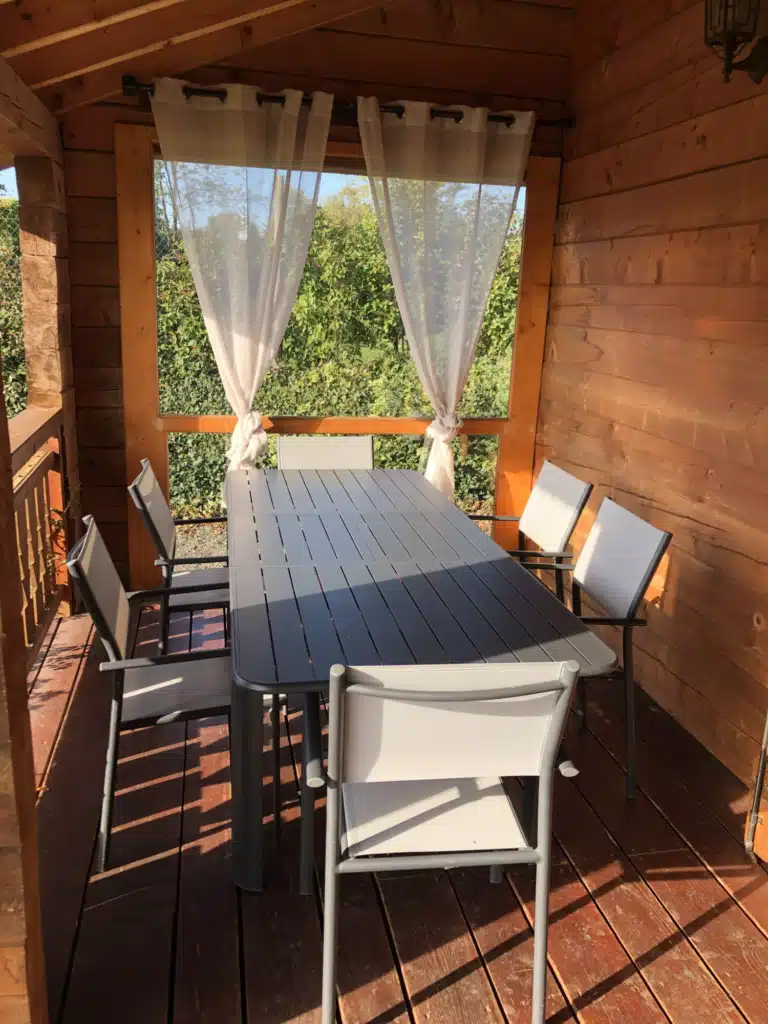 Salle à manger d'extérieur dans une tonnelle en bois : grande table anthracite, 6 chaises claires et rideaux voilés offrant une vue sur le jardin.