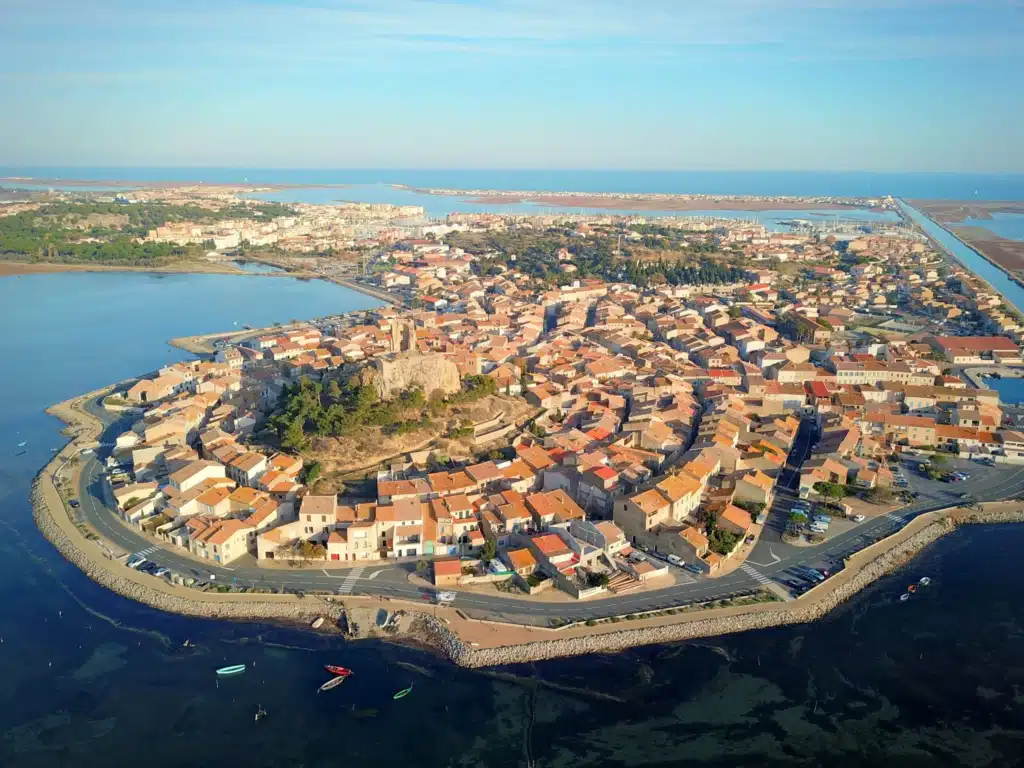 Village méditerranéen en presqu'île : vue aérienne et toits de tuiles. Village historique en presqu'île sur la Méditerranée. Toits de tuiles, maisons en cercle autour d'un rocher fortifié, entre étang et mer.